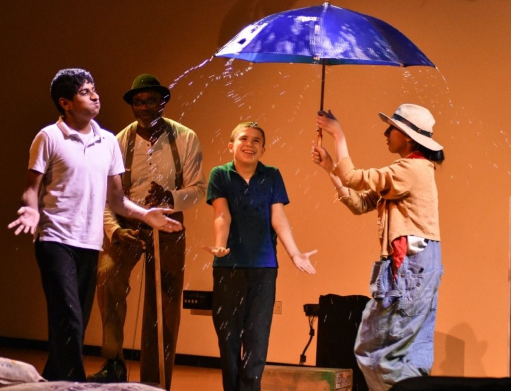 Rain splashes over the top of a blue umbrella twirled by an actor. Two young audience members feel the water spray. A musician plays a wash tub bass in the background.