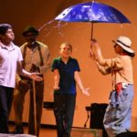 Rain splashes over the top of a blue umbrella twirled by an actor. Two young audience members feel the water spray. A musician plays a wash tub bass in the background.