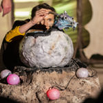 Photo of Bamboozle's PMLD production Makara and the Mountain Dragon. A baby dragon is hatching out of a large egg. His small head peeps over the top as he emerges.