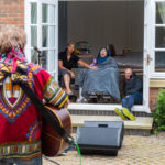 Photo of young person with their Mum and Dad enjoying live music together in their garden through an open double doors. Bamboozle musician is playing a guitar in the foreground.