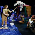 Photo shows a Bamboozle performers with a large puppet of a flying white goose. The goose flies towards a family group who are sitting at the edge of the blue fabric river.
