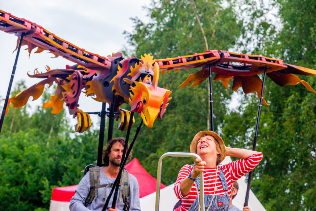 The photo shows the large phoenix puppet flying above the caretaker who stands on a ladder. The Firebird is orange of yellow with wings outspread. The caretaker wears dungarees and a red and white striped long-sleeved t-shirt and looks amazed to see the large phoenix appear.