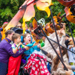 The photos shows the huge Firebird puppet who is swopping down to greet a young girl - she is using a wheelchair and sits with a bright red blanket on her knee. She smiles broadly towards the puppet who comes in close to her. A young boy wearing a t-shirt and red dungarees reaches how to touch the Firebird.