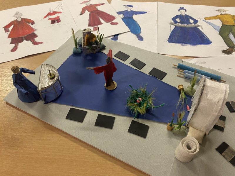 The image depicts designer Nettie Scriven's concept designed for Bamboozle's show The River. This includes a model of the set featuring a bright blue floor cloth with acts as a river and drawing of flowing costumes of bright blues and reds.