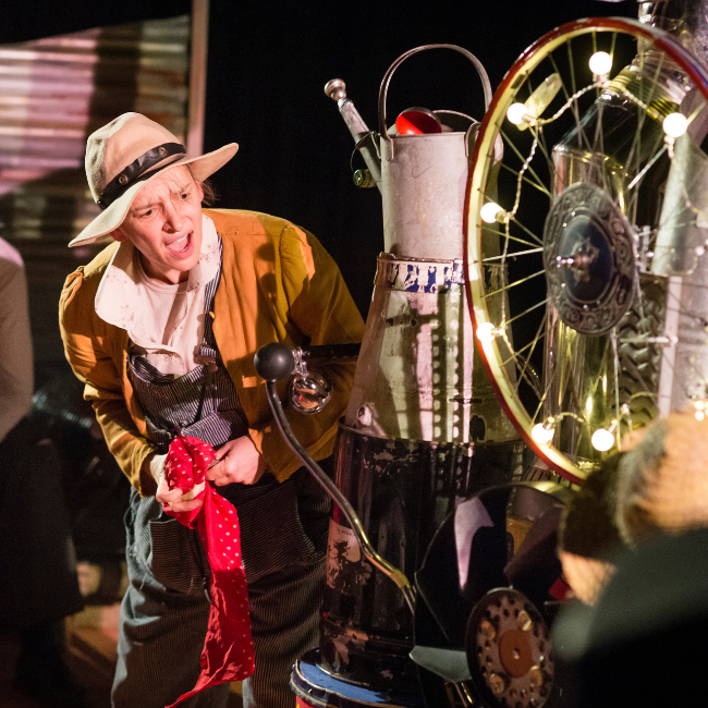 The image shows Clanky Jane who wears a straw hat, denim dungarees and an earthy yellow cardigan. She stands next to her Cloud-O-Matic machine which is made out of an assortment of recycled items including an old watering can, a buggy and a bicycle wheel covered in lights.
