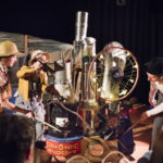 The image shows the character in Pulse investigating Clanky Jane's Cloud-O-Matic machine. The machine is made from an assortment of found items like metal tubes, a bicycle wheel covered in lights, an oil drum and a buggy.