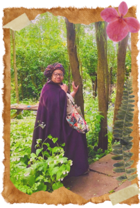 Glendolin is stood in the centre of a photo wearing a dark purple cloak and hat. She is holding a multicoloured patchwork bag. She is stood in a green, woody area full of lush plants and foliage. The photo has ripped edges and looks like it's from a scrapbook page with some pressed plants on top and masking tape on the edges. It has a vintage feel. 