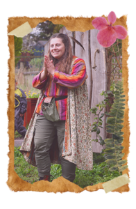 Little Wing is stood by the Backyard gates wearing a floral cloak, brightly coloured striped t-shirt and green trousers. She has long brown hair with a feather in. She is holding her hands up in a praying position and smiling at a family off camera.