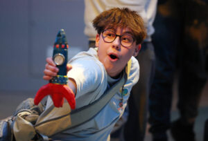 Lou is performing in Moon Song holding a rocket out in front of them with an awed expression. Photo by Darren Staples.