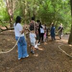 A group of families and artists are walking through the woods all holding onto a long piece of rope and wearing headphones