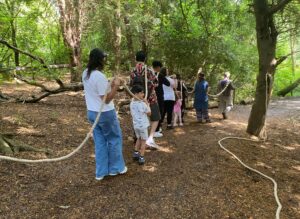 A group of families and artists are walking through the woods all holding onto a long piece of rope and wearing headphones