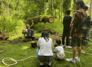 A group of families are gathered in the woods watching a Bamboozle artist play accordion